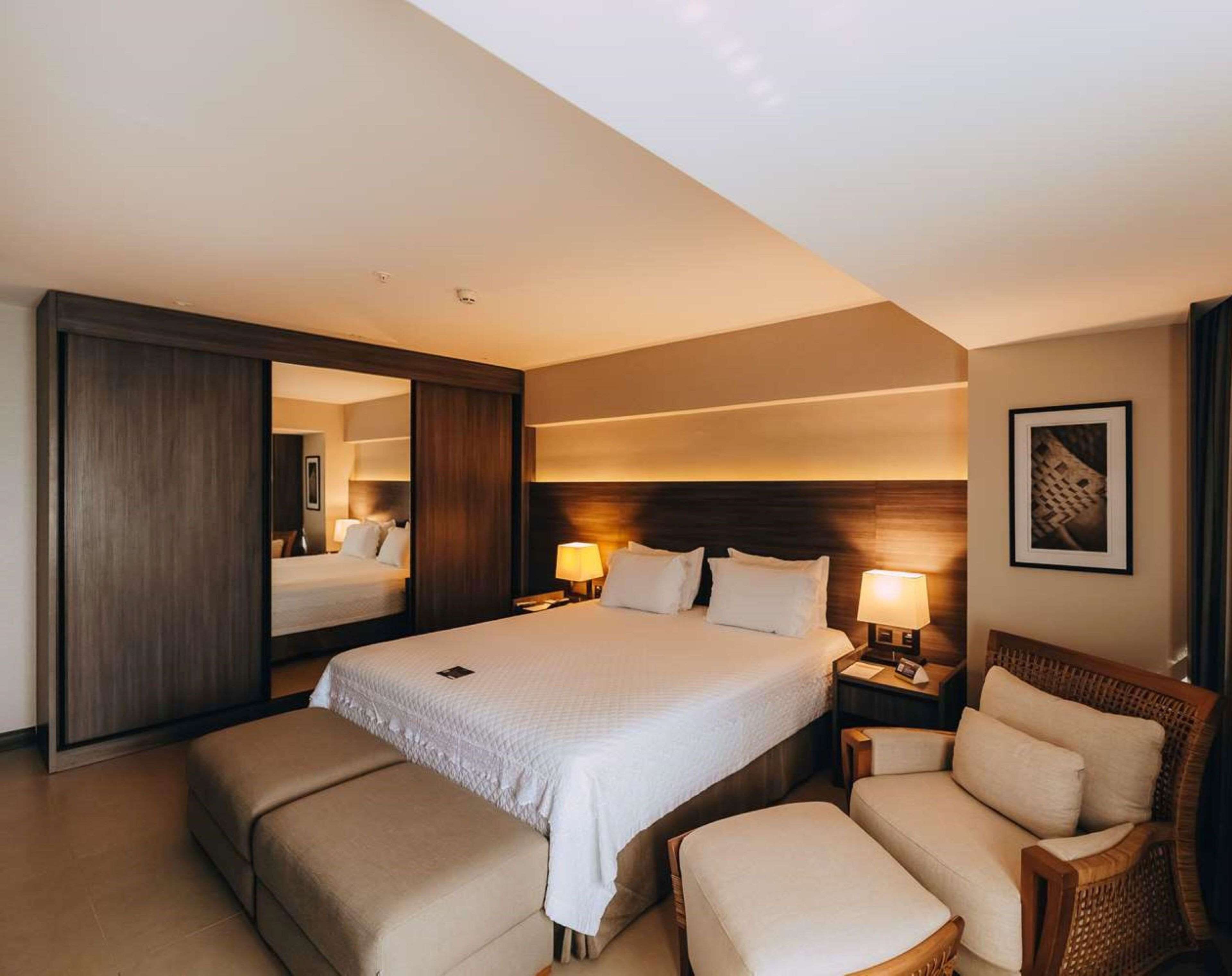 Una habitación de hotel con una cama grande, una zona de estar con una silla y reposapiés, una mesita de noche con lámpara y un espejo.