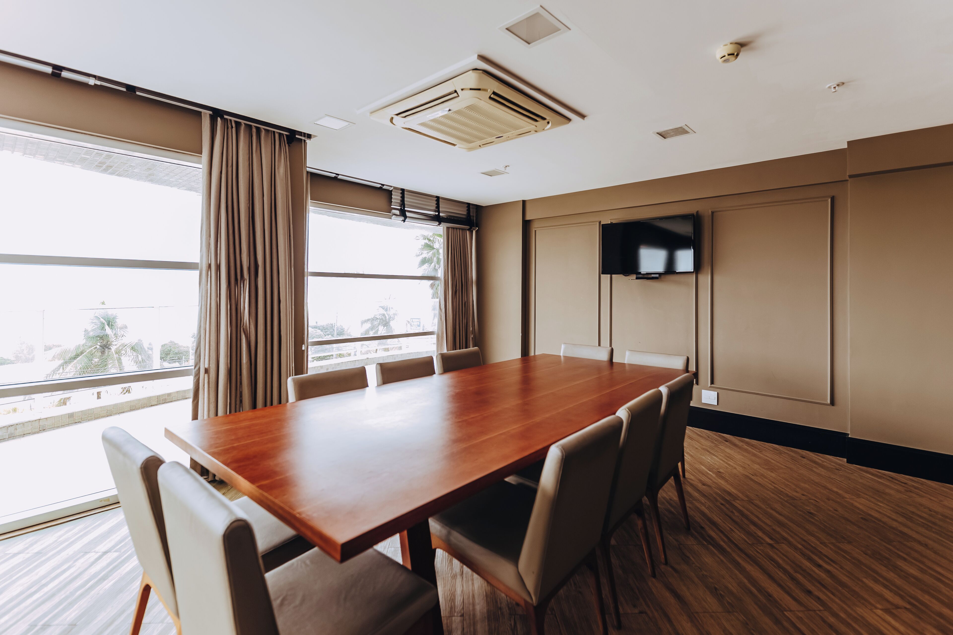 A conference room with a wooden table, beige chairs, a large window with curtains, a mounted TV, and an air conditioning unit.