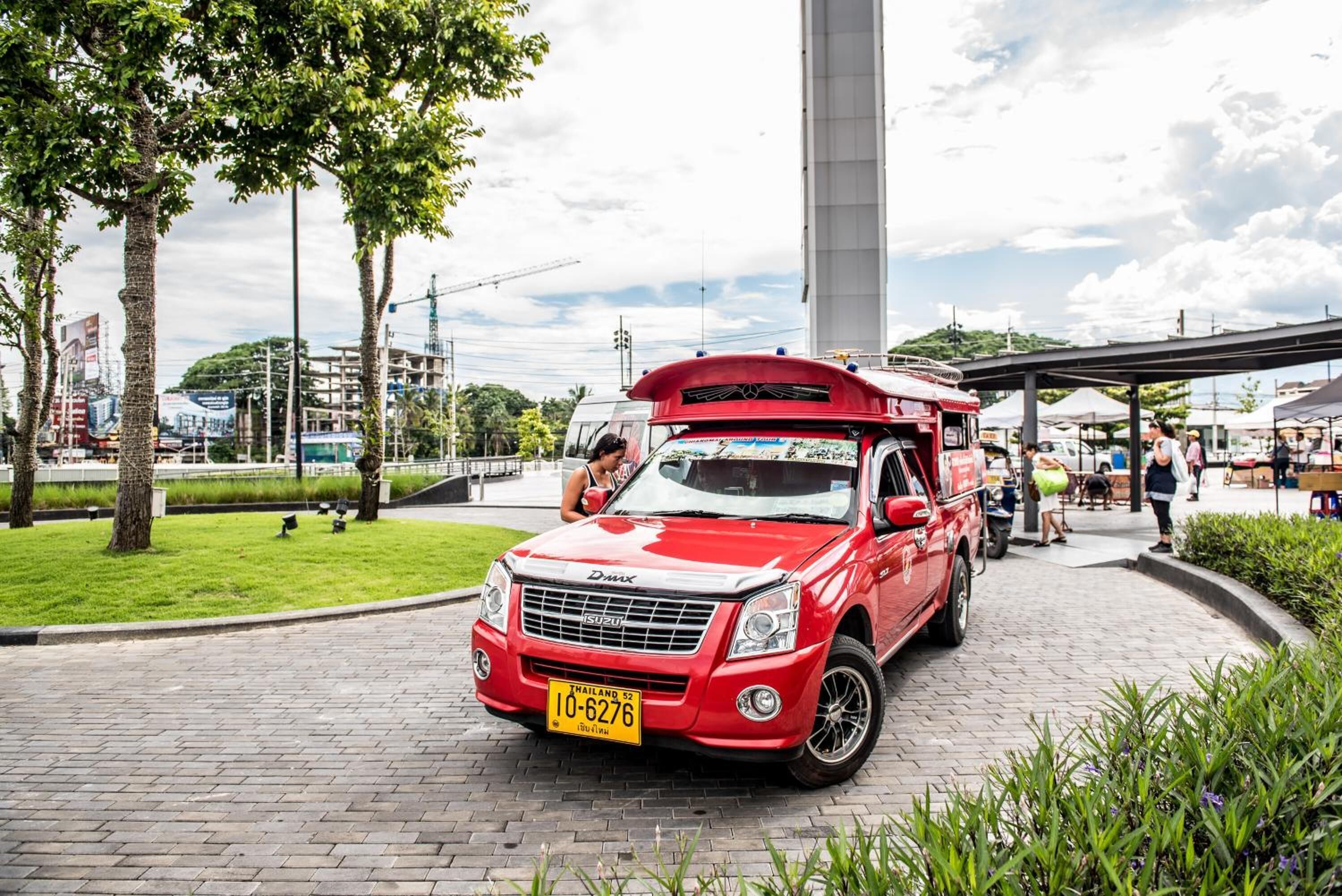 A red Suzuki D-Max truck parked on a paved area with a yellow license plate, surrounded by greenery and a pedestrian walkway.