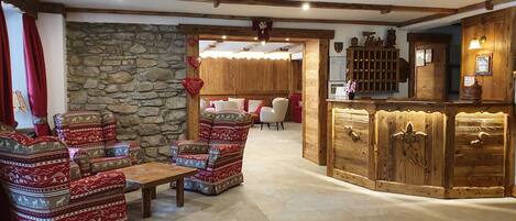 Un lobby acogedor con sillones estampados, pared de piedra, vigas de madera y una mesa de recepción.