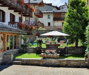 Un patio con asientos al aire libre, paredes de piedra y una escalera de madera que conduce a un balcón con jardineras.