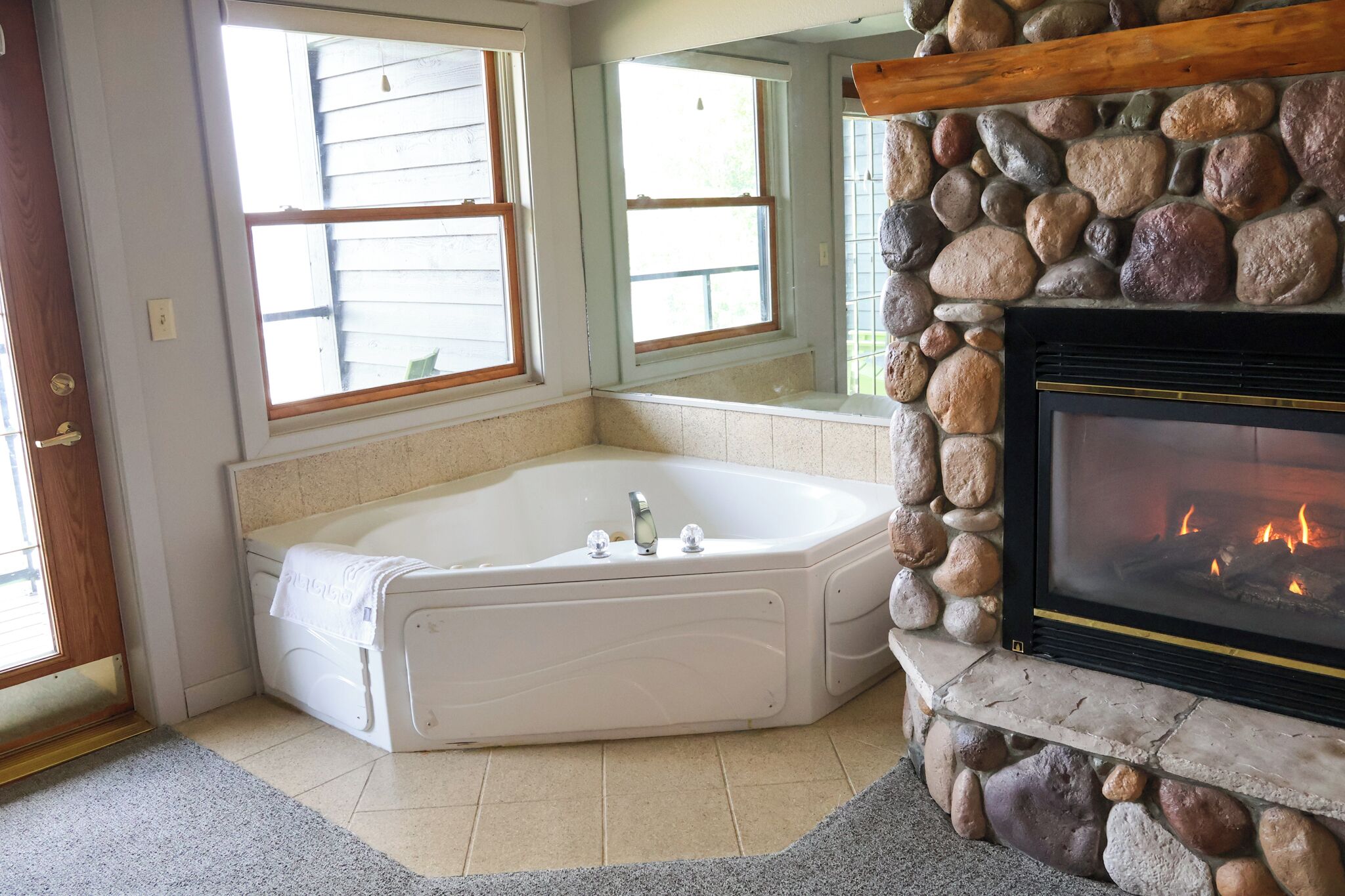A corner bathroom with a large bathtub, a stone fireplace, and a window with wooden shutters.