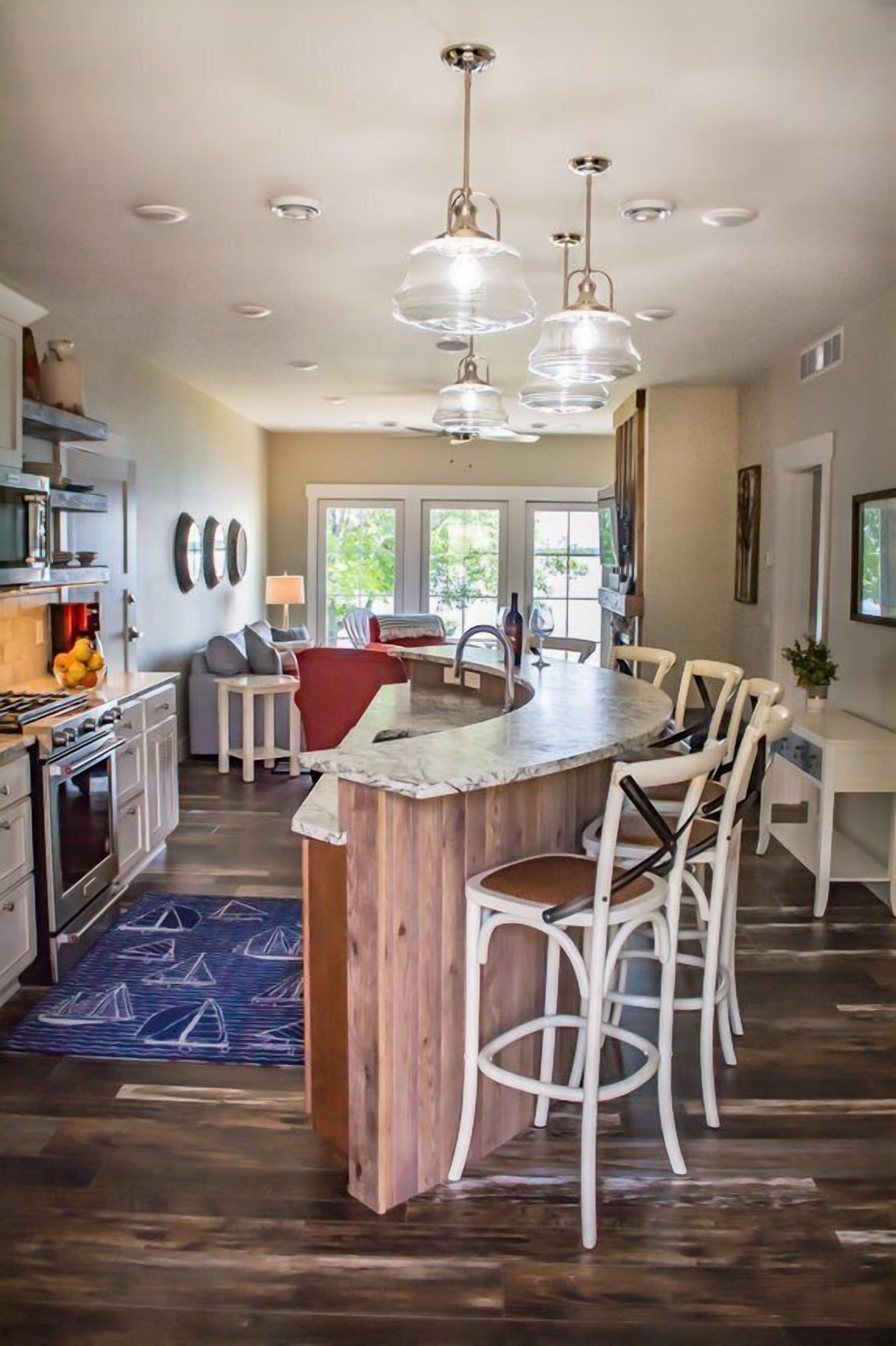 A modern kitchen with a central island, white bar stools, pendant lighting, and a view of the outdoors.