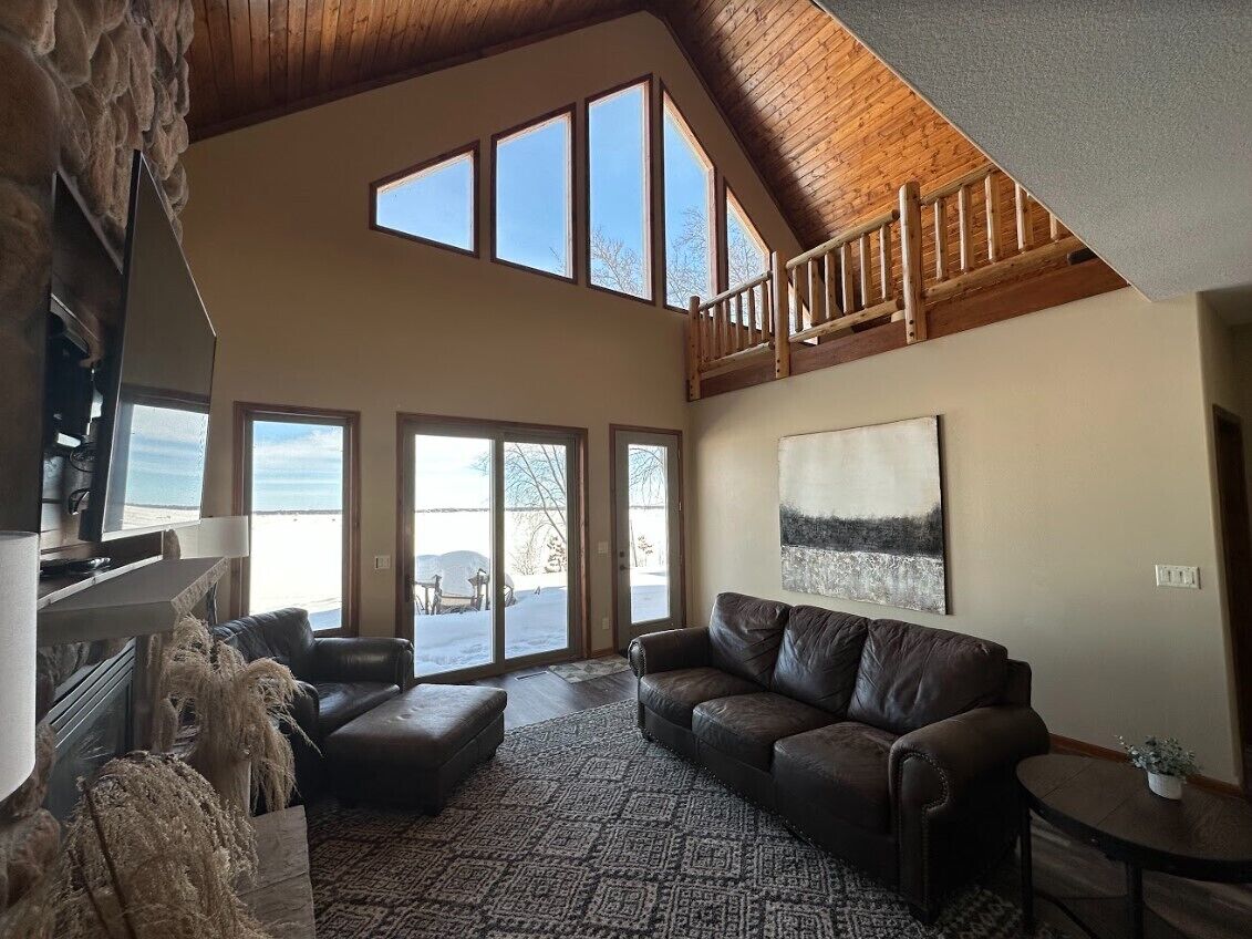 A spacious living area with a high ceiling, a stone fireplace, a large window, a brown leather sofa, and a painting on the wall.
