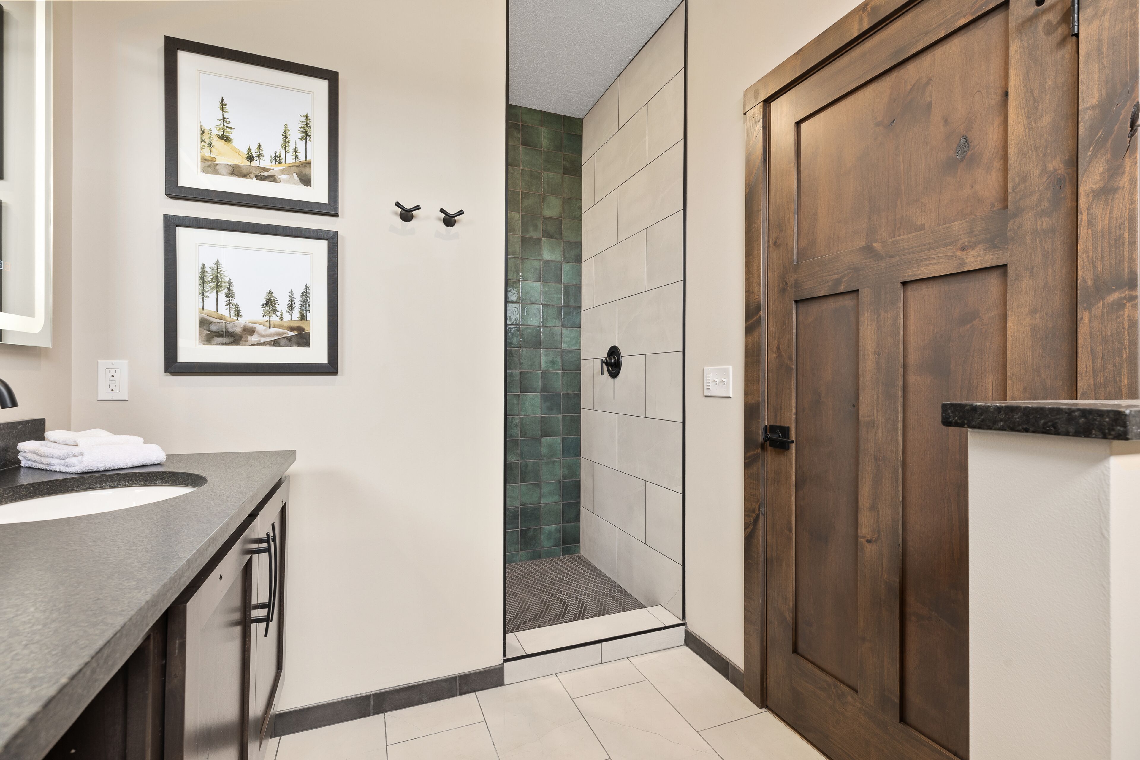 A bathroom with a wooden door, a sink with a dark countertop, and two framed pictures on the wall.