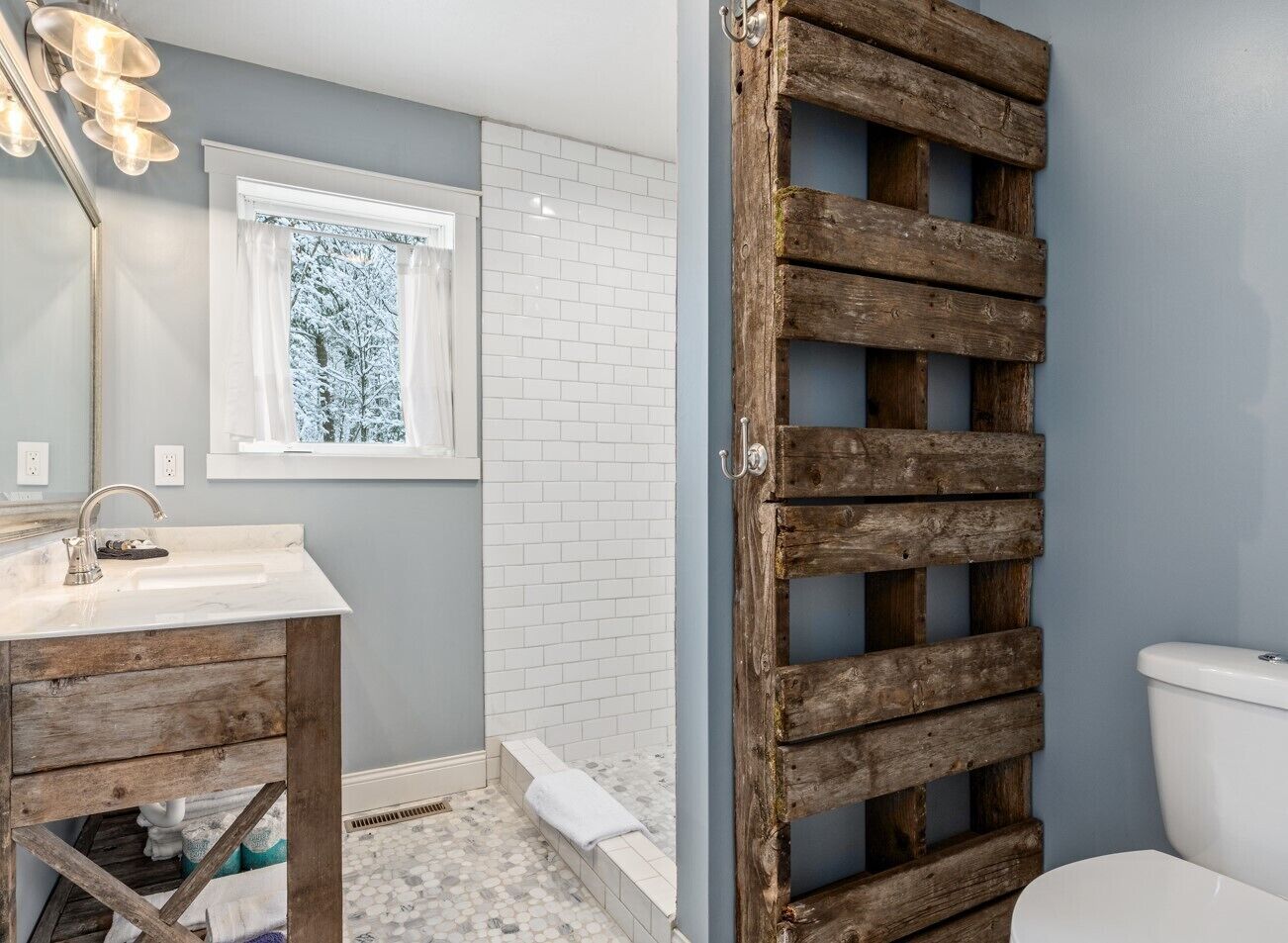 A bathroom with a wooden vanity, a white sink, a shower with a white tiled wall, and a rustic wooden door.
