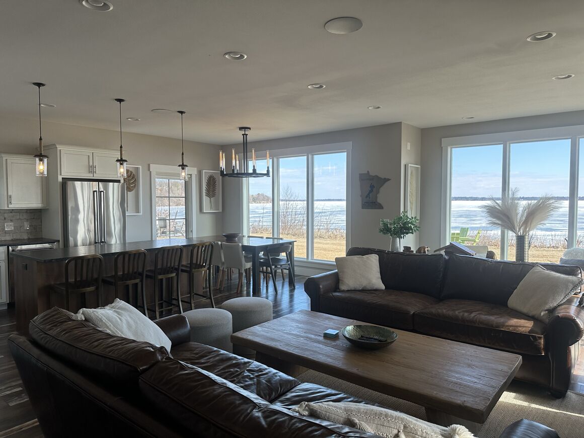 A spacious living area with a dark brown leather sofa, a wooden coffee table, and a dark brown couch. A kitchen area with white cabinets and a stainless steel refrigerator is visible in the background.