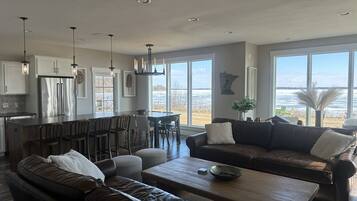 A spacious living area with a dark brown leather sofa, a wooden coffee table, and a dark brown couch. A kitchen area with white cabinets and a stainless steel refrigerator is visible in the background.