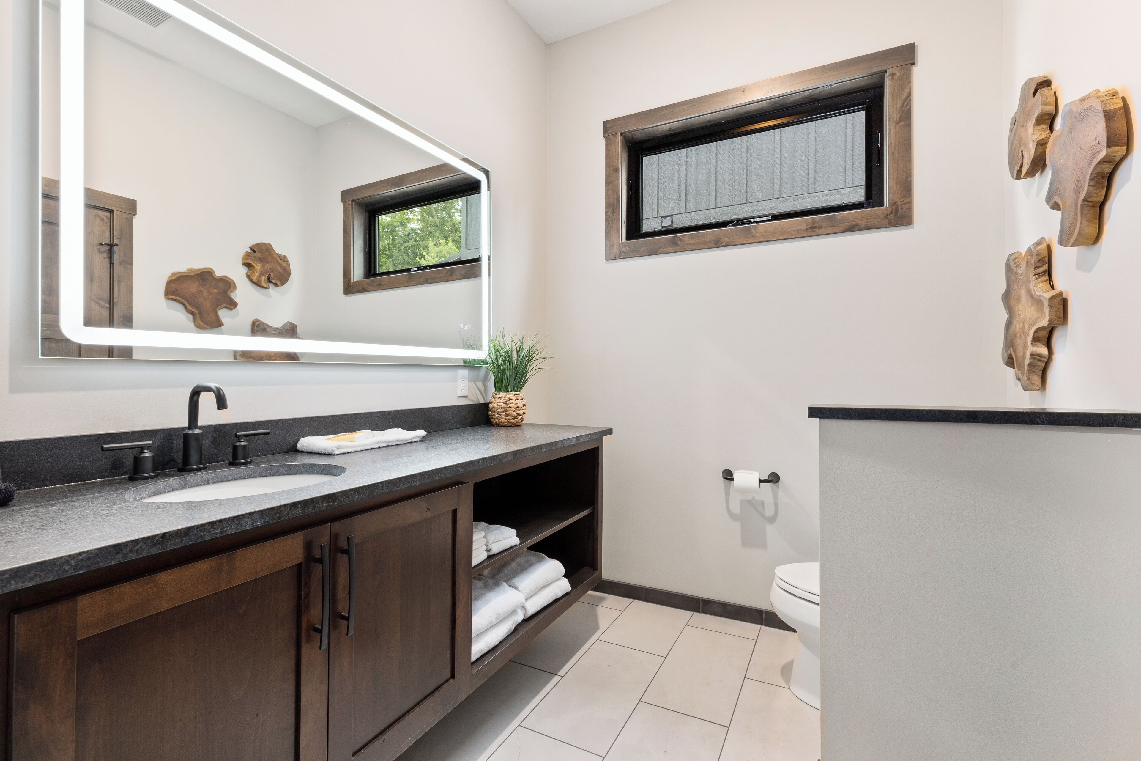 A modern bathroom with a large mirror, dark wood cabinets, a granite countertop, and a window above the toilet.