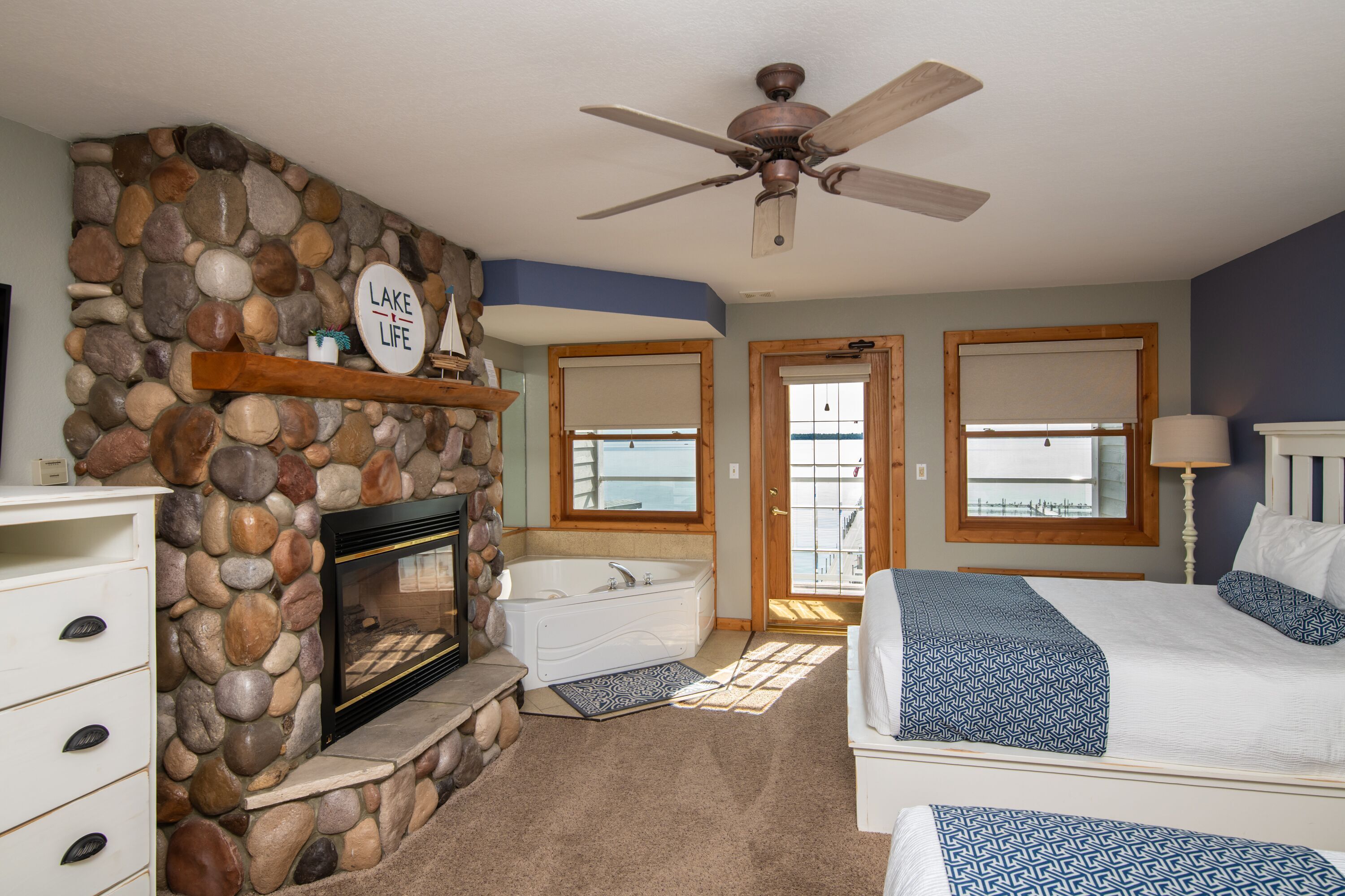 A bedroom with a stone fireplace, a bathtub, a bed with patterned bedding, and a ceiling fan.
