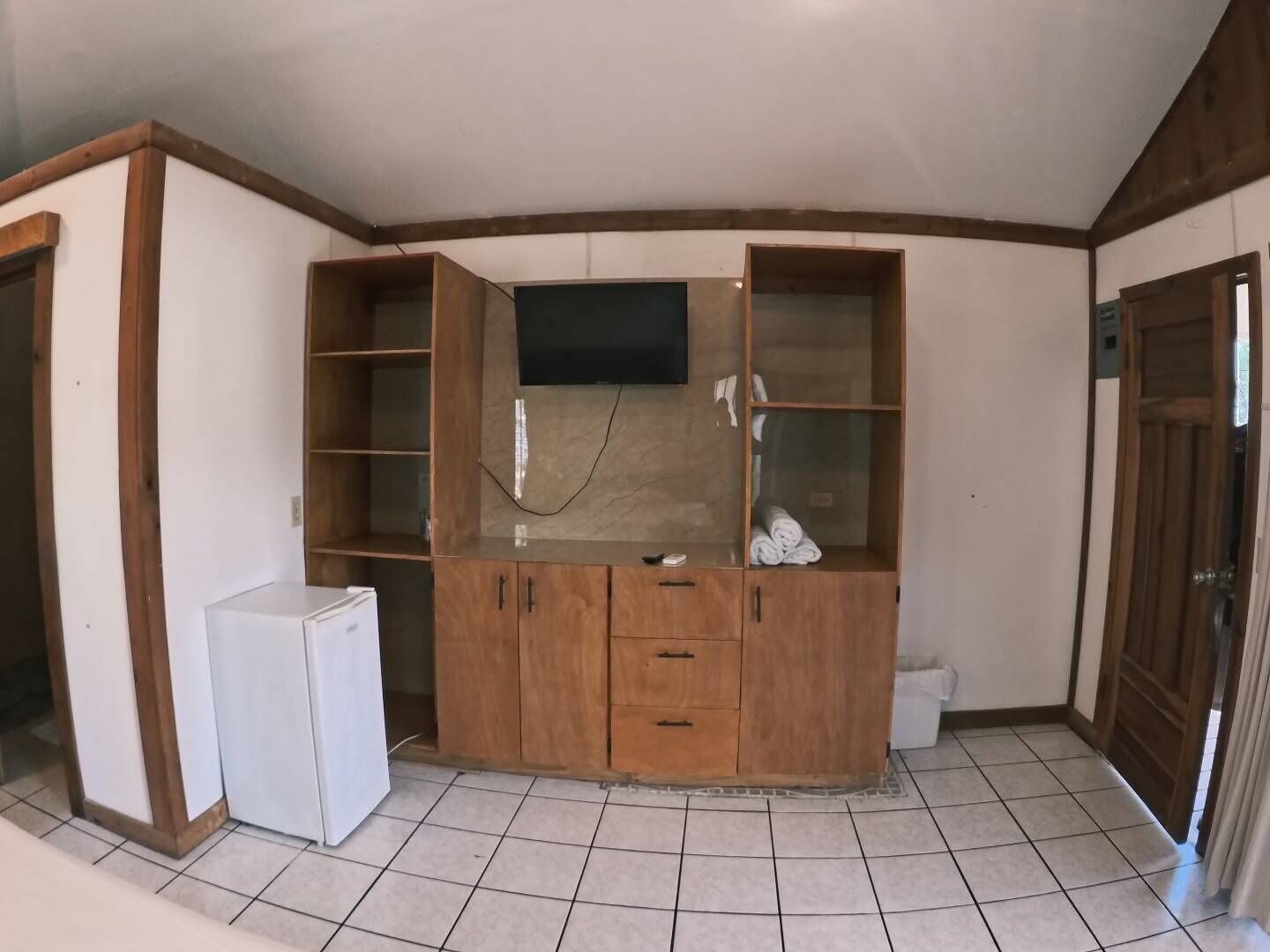 A room with a built-in wooden cabinet with a flat-screen TV, a small refrigerator, and a tiled floor.