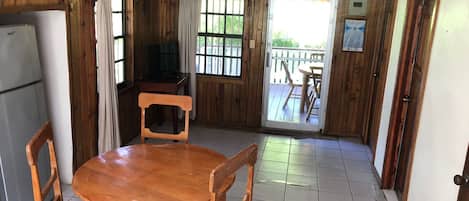 A wooden interior with a round dining table, chairs, a refrigerator, and a sliding glass door leading to an outdoor area.