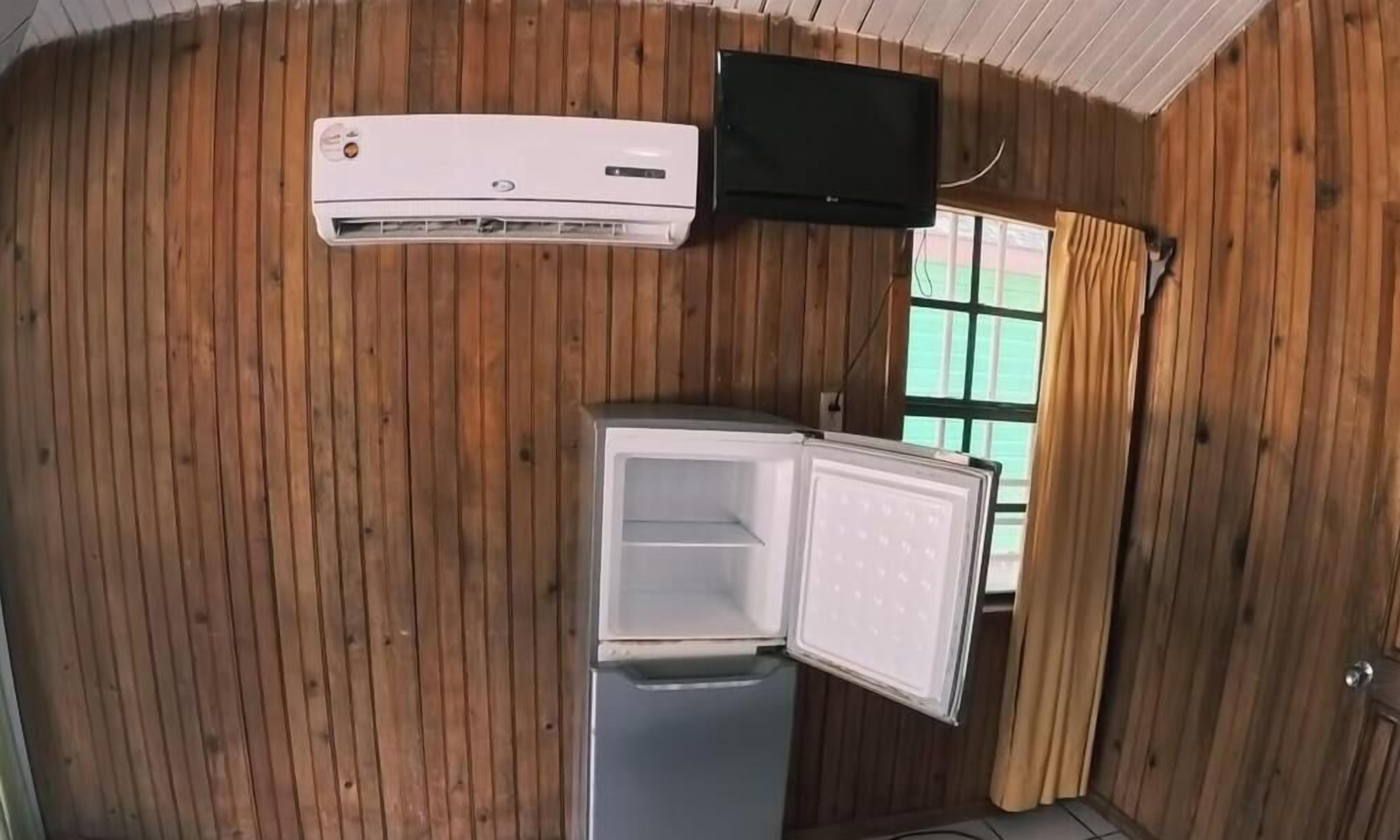 A compact room with wooden paneling, a white air conditioning unit, a television mounted on the wall, and a small refrigerator.