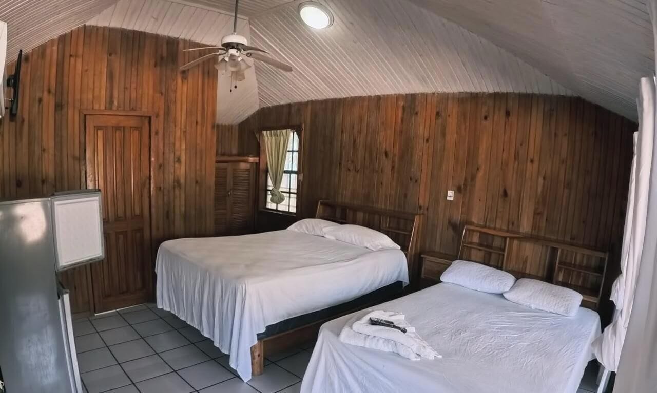 Ett rum med två sängar, träväggar, en takfläkt och ett fönster med gardiner.