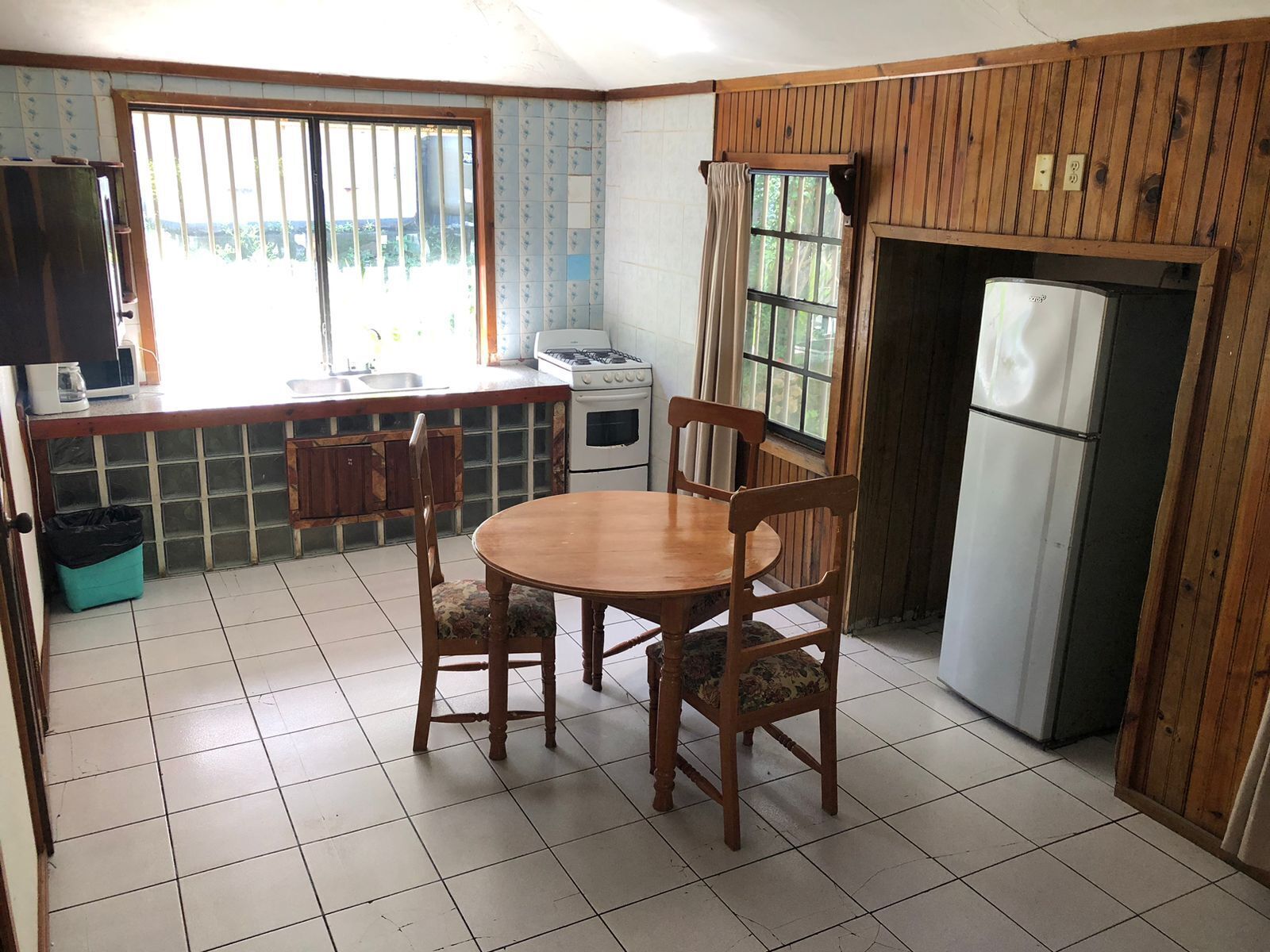 Une cuisine avec une table et des chaises en bois, un fourneau, un réfrigérateur et une fenêtre avec des barreaux.