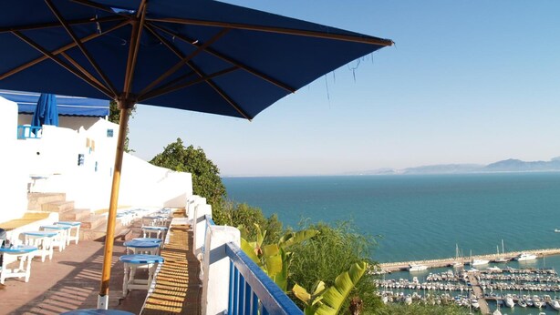 Une terrasse agrémentée de parasols bleus, entourée de bâtiments blancs, et offrant une vue sur la mer et les bateaux.