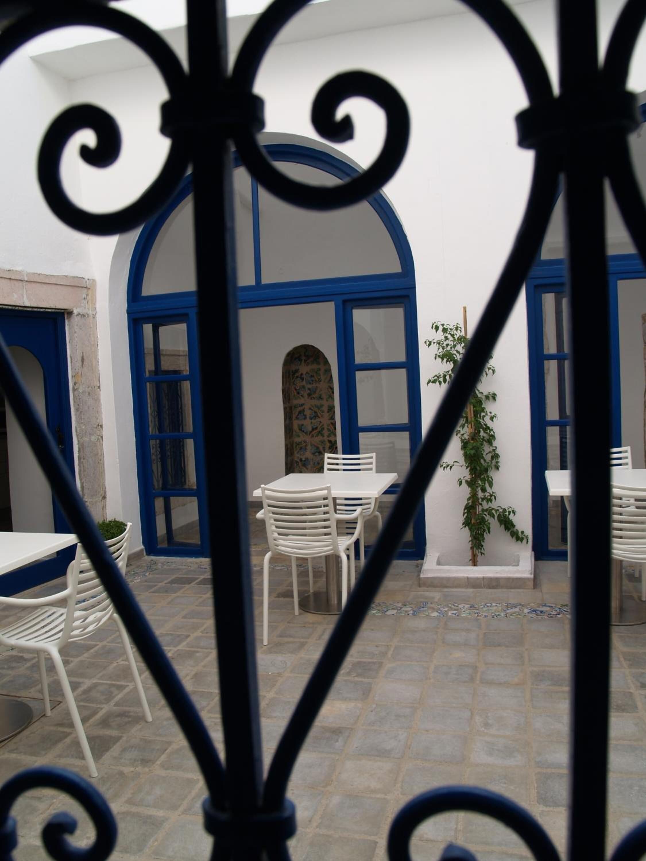 Um pátio com mesas e cadeiras brancas, portas azuis e piso de azulejos.