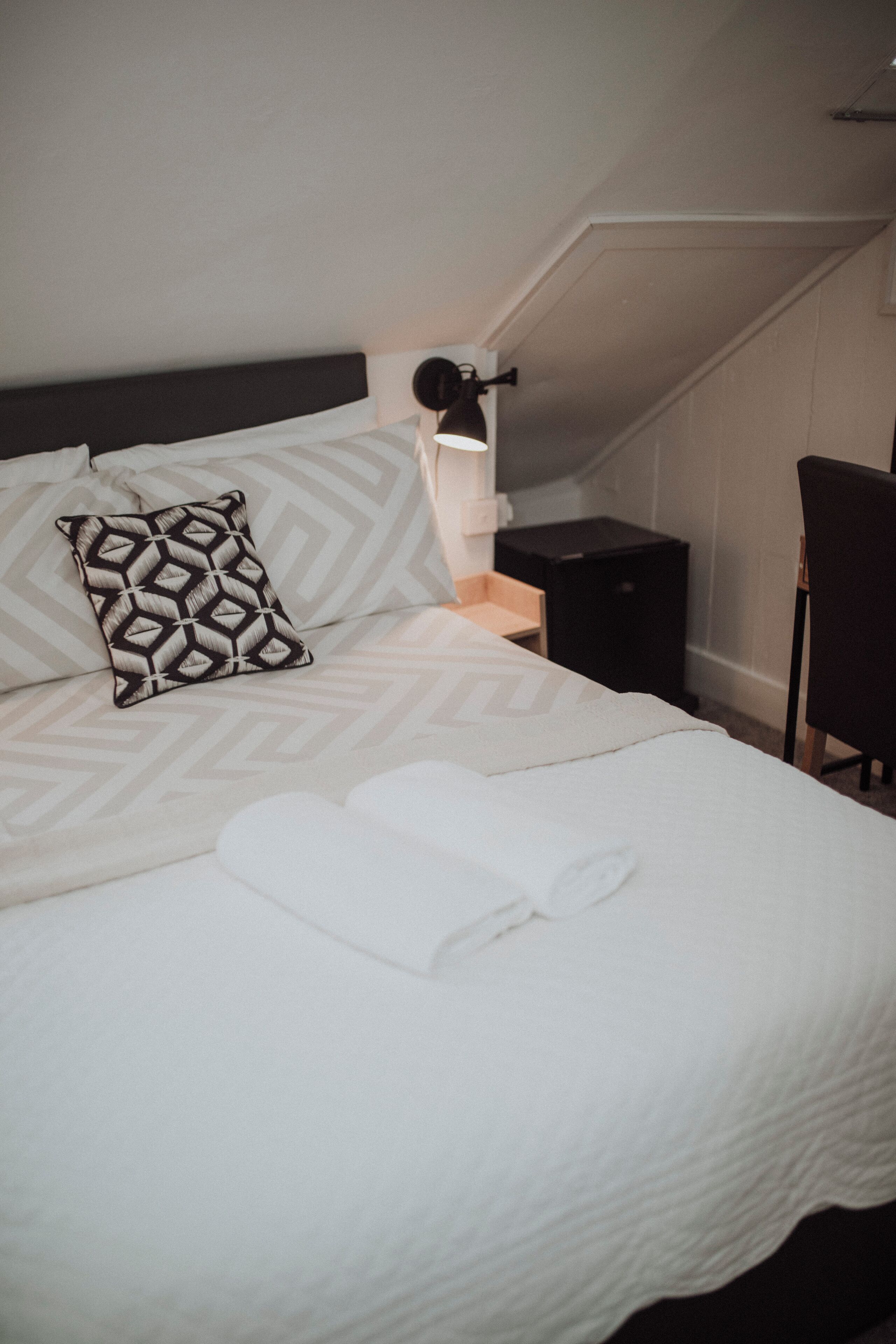 Un lit bien fait, agrémenté d’un coussin à motifs, une table de chevet avec une lampe et une chaise dans une chambre d’hôtel.