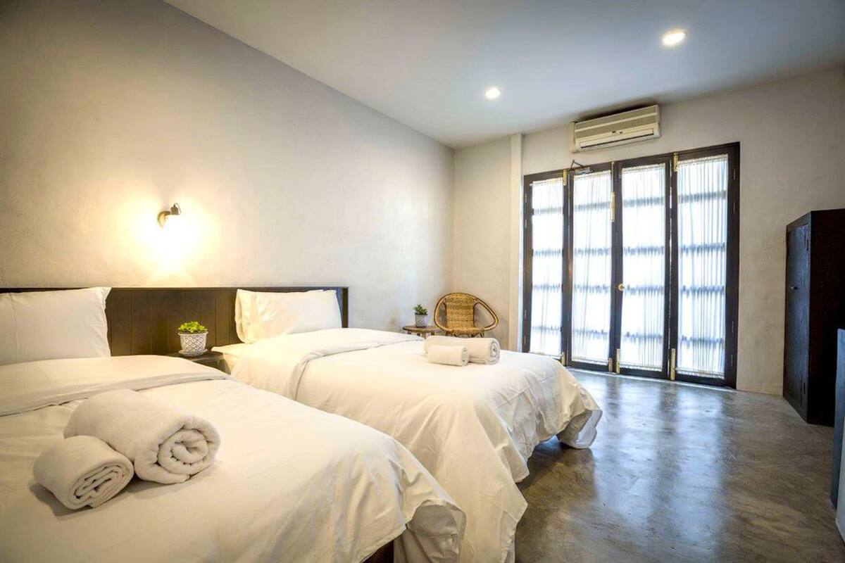 A hotel room with two beds, a small table with a plant, a chair, and a large window with blinds.