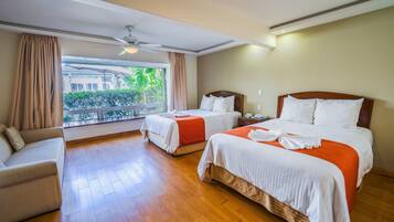 A hotel room with two beds, a sofa, and a balcony with a view of greenery.