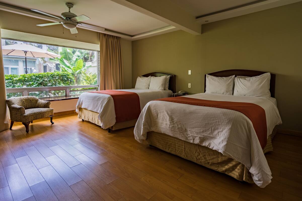 Een hotelkamer met twee bedden, een stoel en een groot raam met uitzicht op groen.