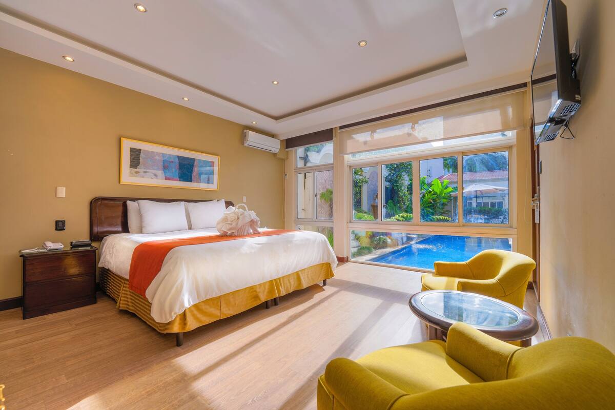 Hotelkamer met een bed, een klein tafeltje, gele stoelen, een televisie en uitzicht op een zwembad.