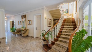 Una imponente escalera con barandillas ornamentales que sube a un segundo piso, un vestíbulo espacioso con piso de mármol y una planta grande en maceta.