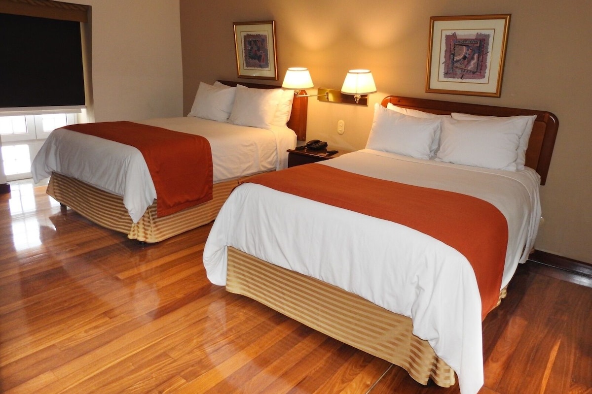 A hotel room with two beds, wooden flooring, and wall-mounted lamps.