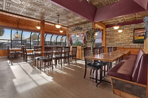 A restaurant with wooden tables and chairs, a stone wall, and large windows. - Guest House Inn & Suites (Edson)