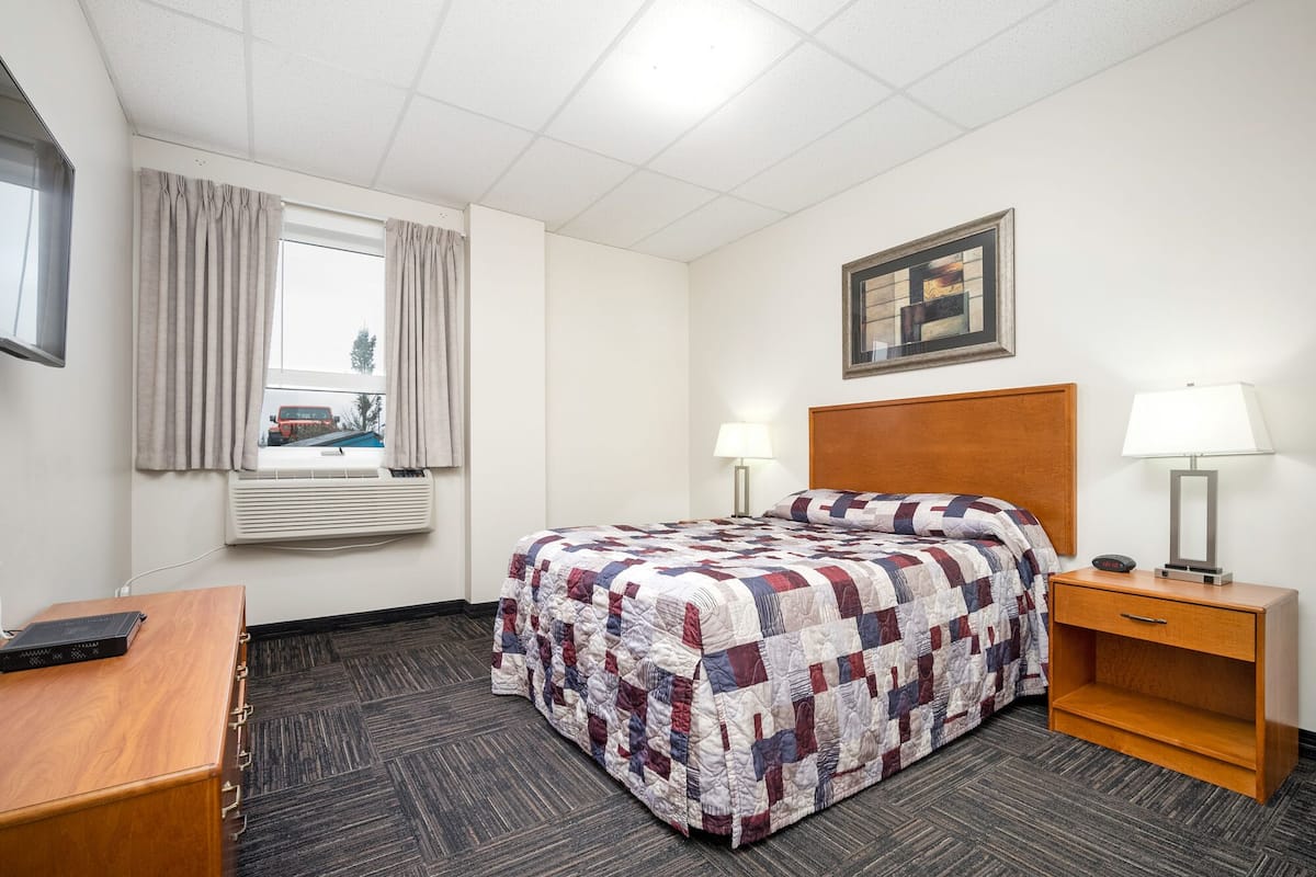 Hotellihuone, jossa on sänky, kaksi yöpöytää valaisimineen, työpöytä, ikkuna verhoineen, ilmastointilaite ja seinällä oleva kehystetty taulu.