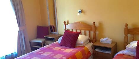 A hotel room with two beds, each with a wooden headboard and a colorful quilt. There is a nightstand with a lamp and a telephone on one side, and a small table with a chair on the other. A mirror hangs on the wall above the beds.