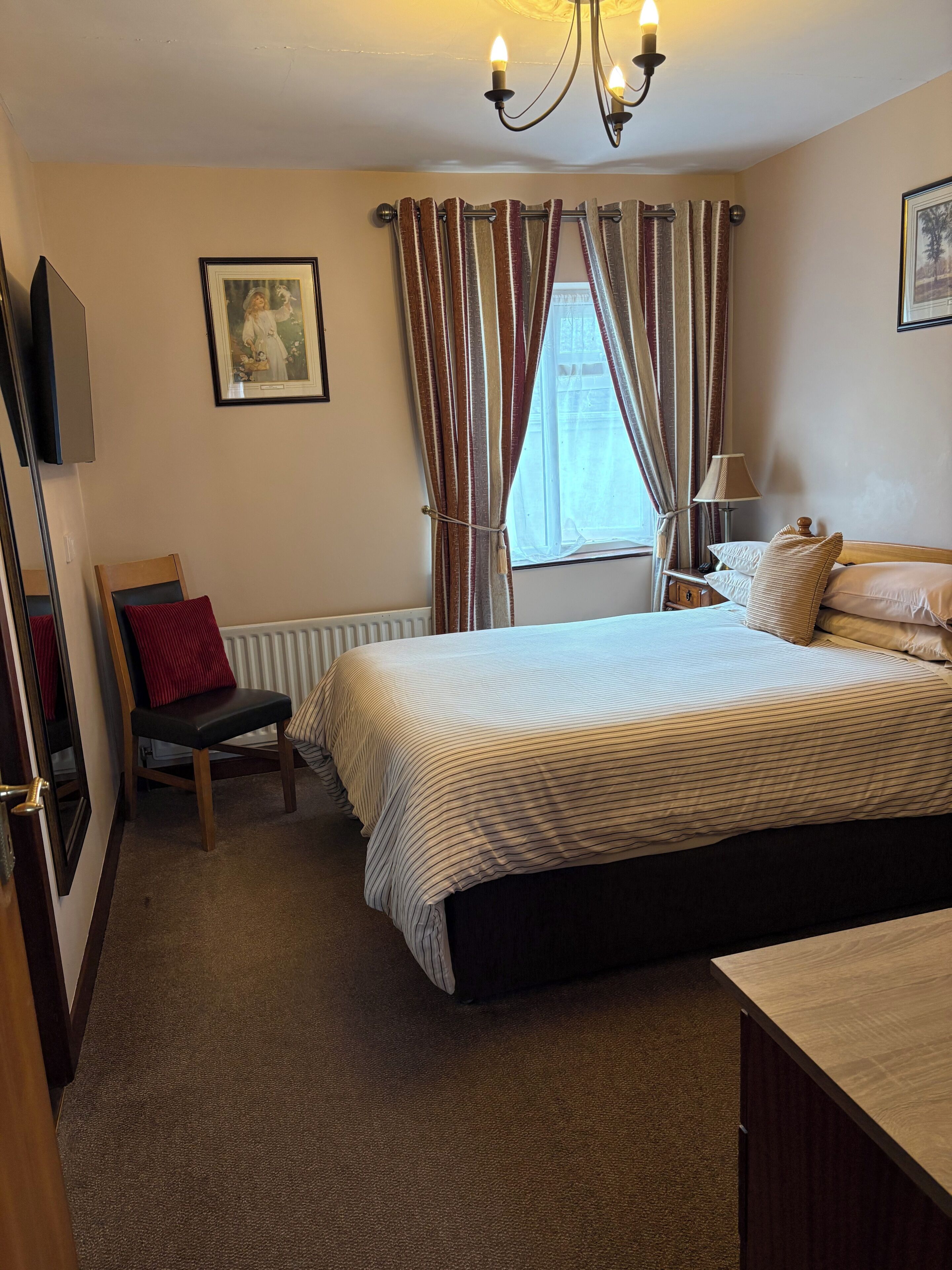 Quarto de hotel com uma cama grande, uma cadeira, uma escrivaninha, um radiador, uma televisão e uma janela com cortinas.