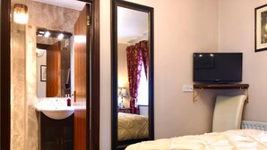 Habitación de hotel con cama, televisión montada en la pared, mesita de noche, espejo y baño con lavabo y ducha.