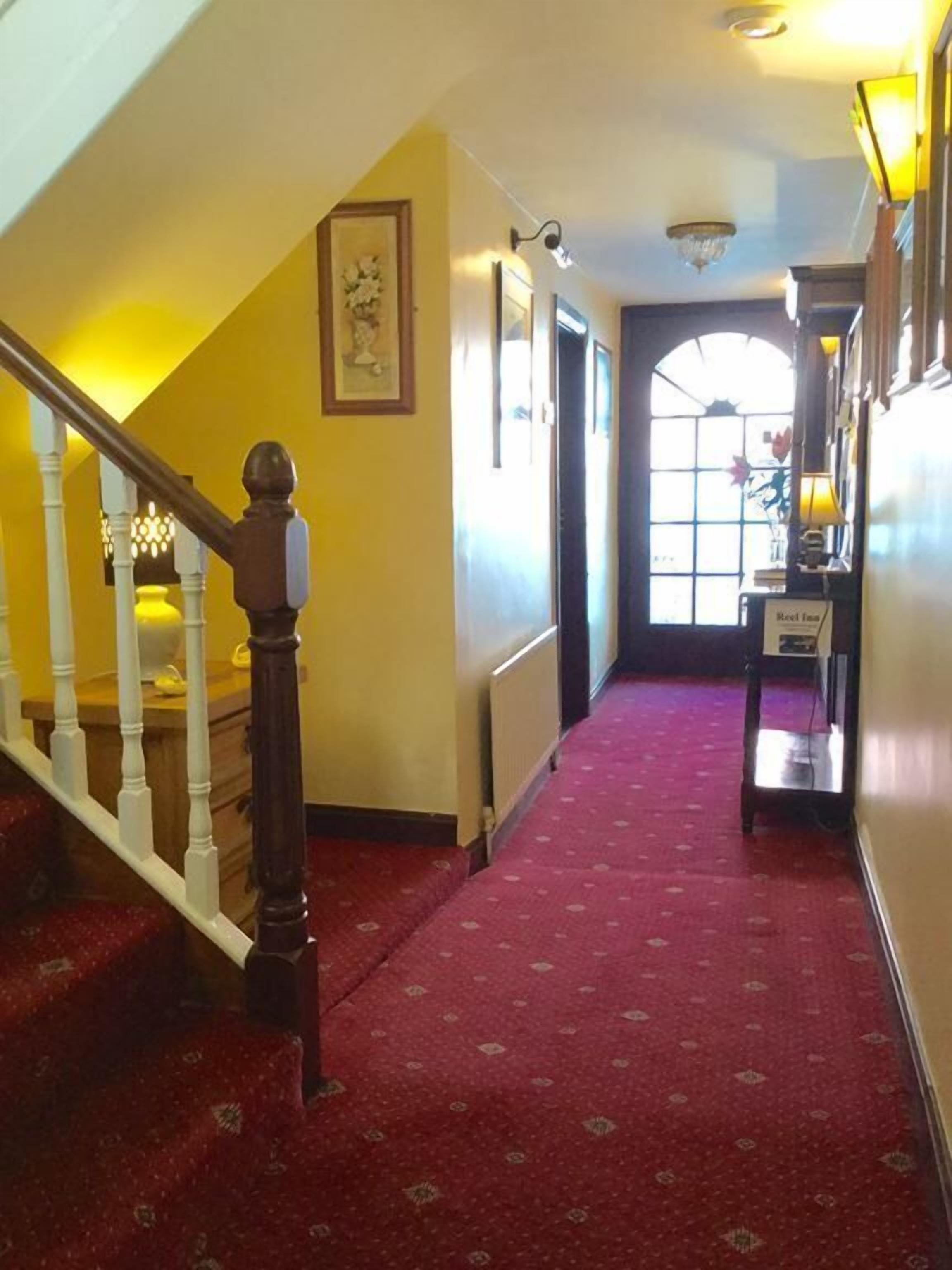 Un pasillo de hotel con alfombra roja, una escalera de madera con balaustres blancos y un candelabro.