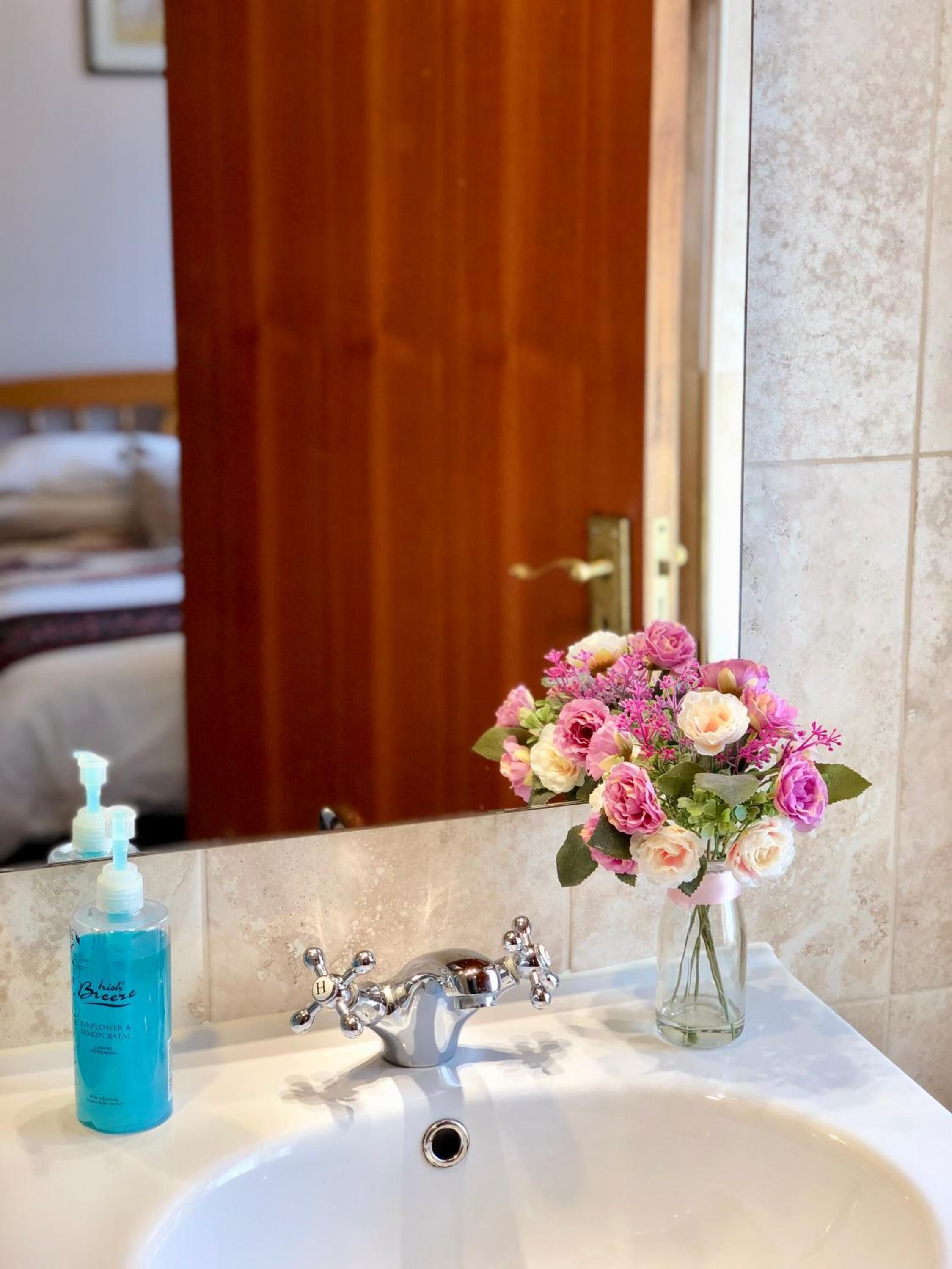 A bathroom with a white sink, a silver faucet, and a mirror reflecting a wooden door and a bed.