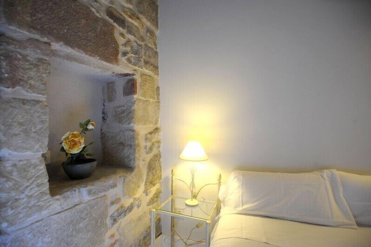A stone wall with a small shelf holding a potted plant. There is a bedside table with a lamp next to a bed.