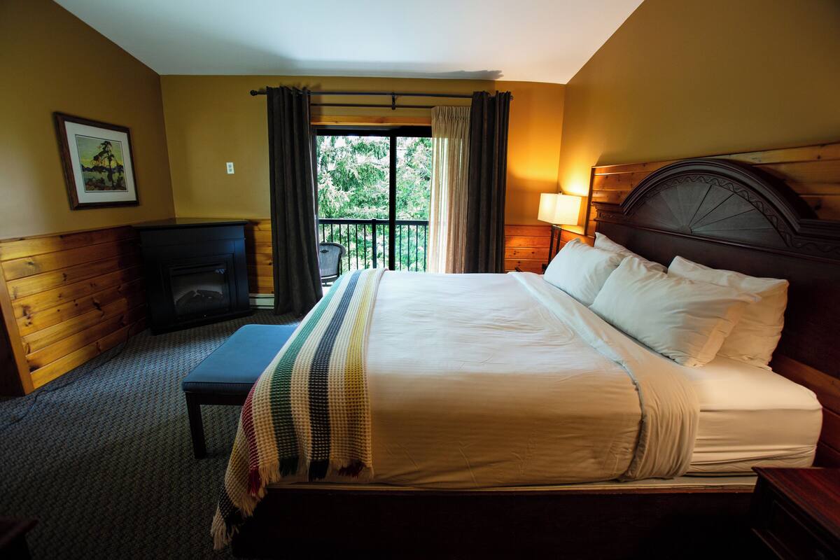Quarto de hotel com uma cama grande, um banco de madeira, uma lareira e varanda com cortinas.