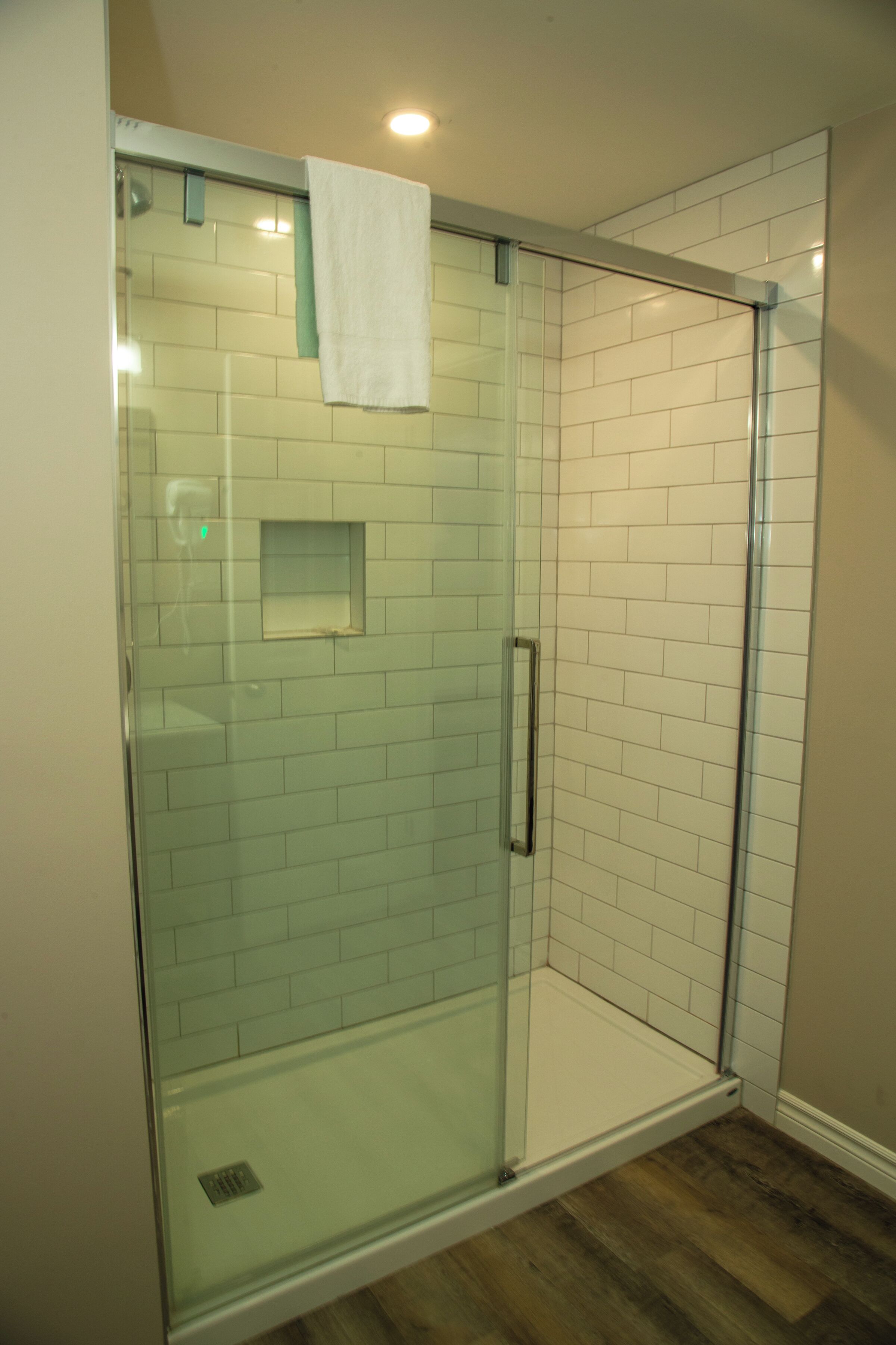 A glass-enclosed shower with white tiles and a wooden floor.