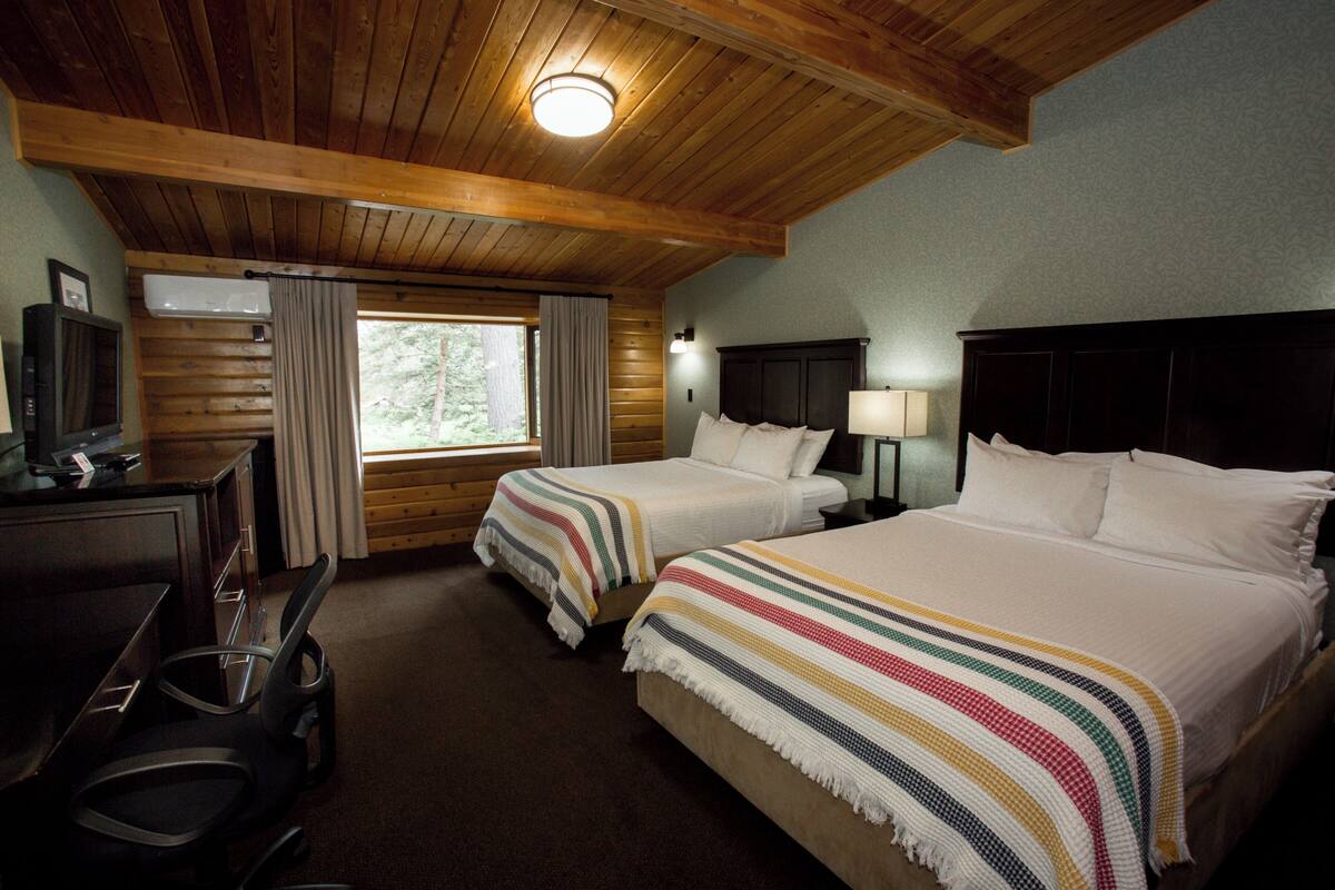 Quarto de hotel com duas camas, uma escrivaninha com cadeira, televisão e uma janela com cortinas.
