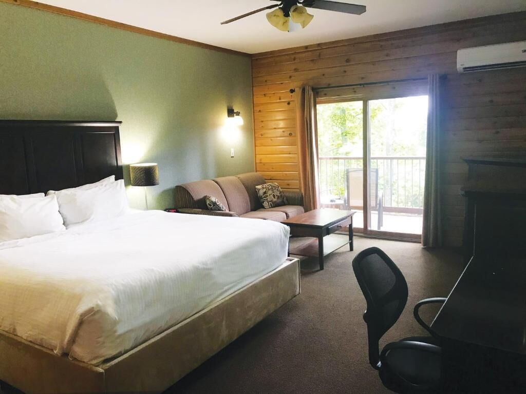Habitación de hotel con una cama grande, un sofá, una mesita, una silla, un ventilador de techo y vistas al exterior a través de un ventanal corredero.