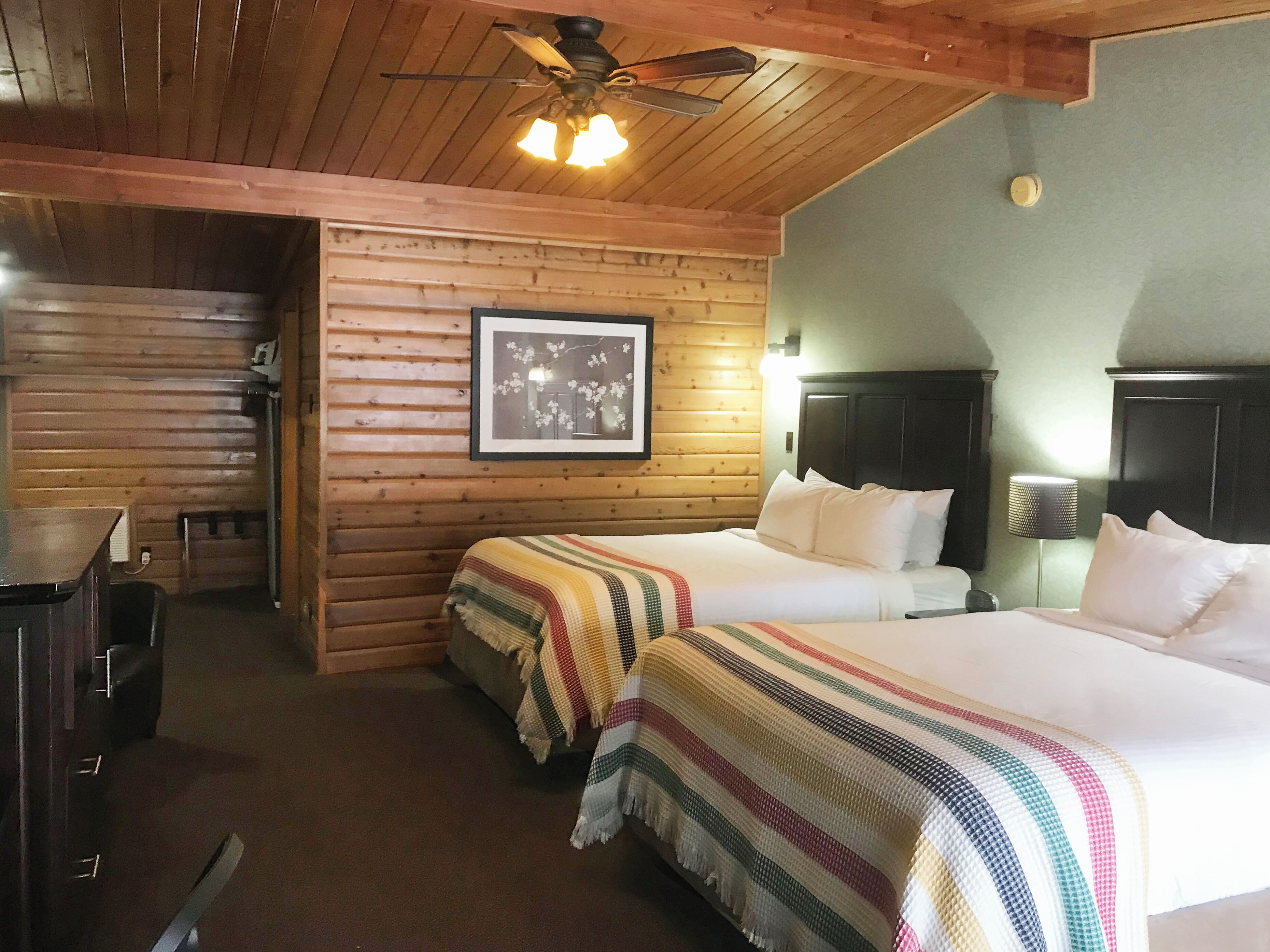 A room with two beds, wooden walls, a ceiling fan, and a framed picture on the wall.
