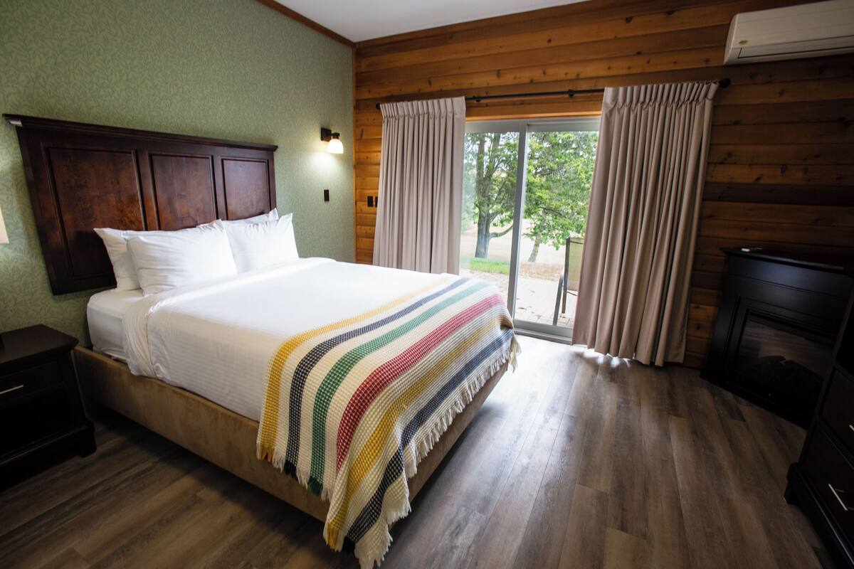 Quarto de hotel com uma cama grande, cabeceira de madeira e vista para o exterior através de uma porta de vidro deslizante.