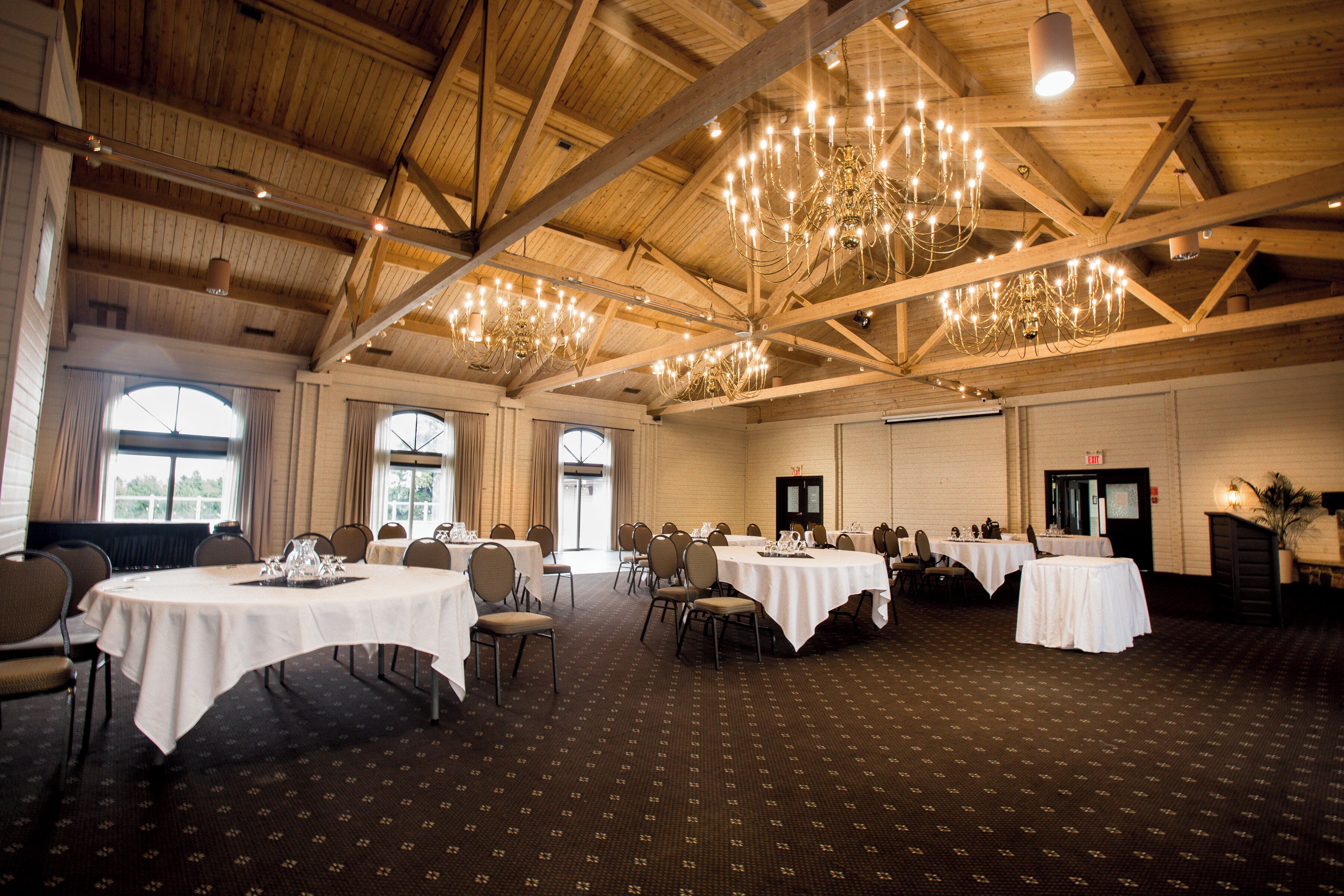 A spacious banquet hall with high ceilings and chandeliers, featuring multiple round tables and chairs, large windows, and a patterned carpet.