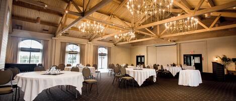 A spacious banquet hall with high ceilings and chandeliers, featuring multiple round tables and chairs, large windows, and a patterned carpet.