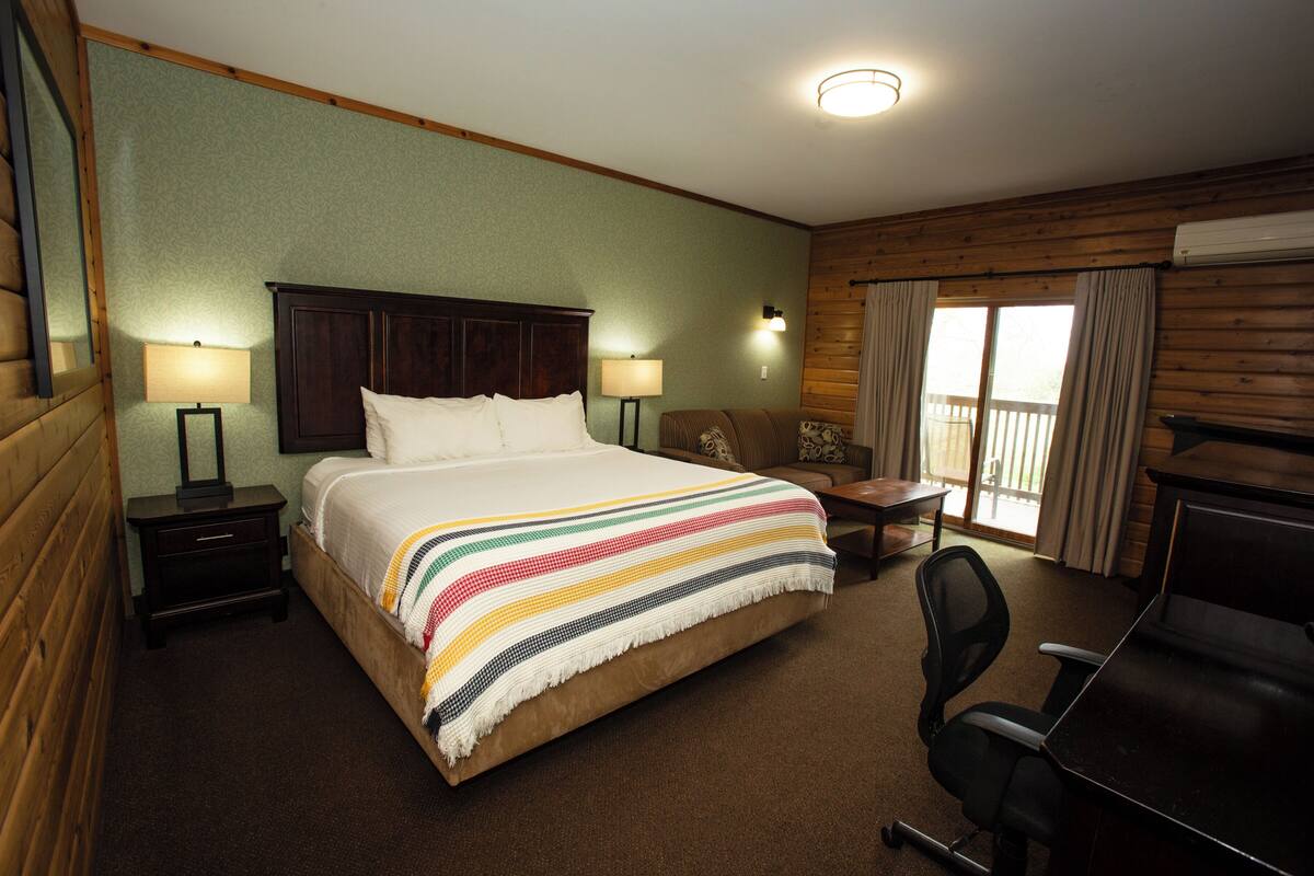 Quarto de hotel com uma cama grande, duas mesas de cabeceira com luminárias, uma escrivaninha com cadeira e vista para o exterior através de uma janela com cortinas.