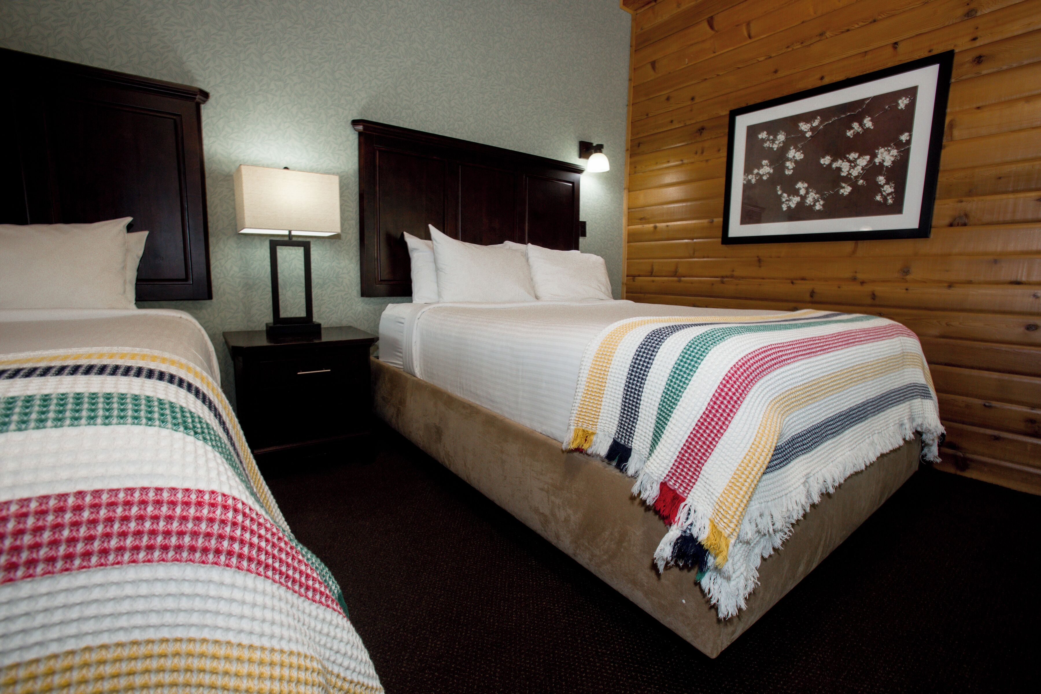 Habitación de hotel con dos camas, cabeceros de madera, una mesita de noche con lámpara y un cuadro en la pared.