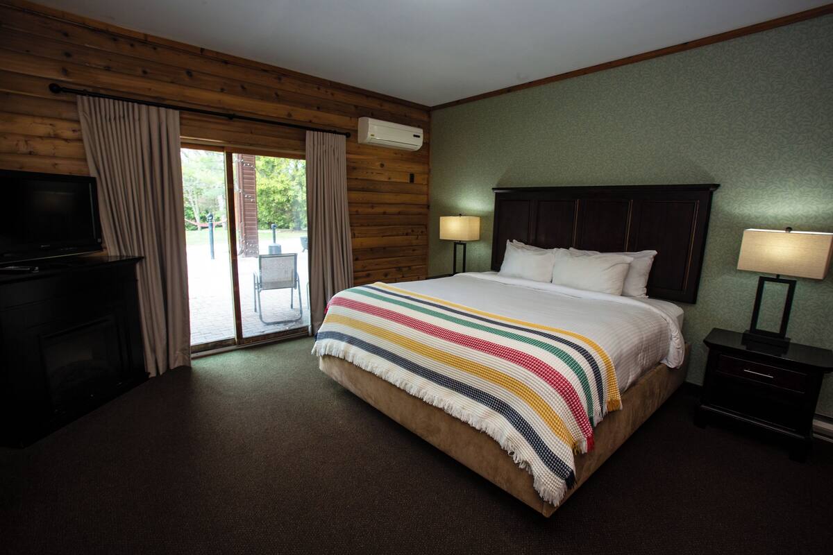 Quarto de hotel com uma cama grande, duas mesas de cabeceira com luminárias, uma televisão e uma porta de vidro que se abre.