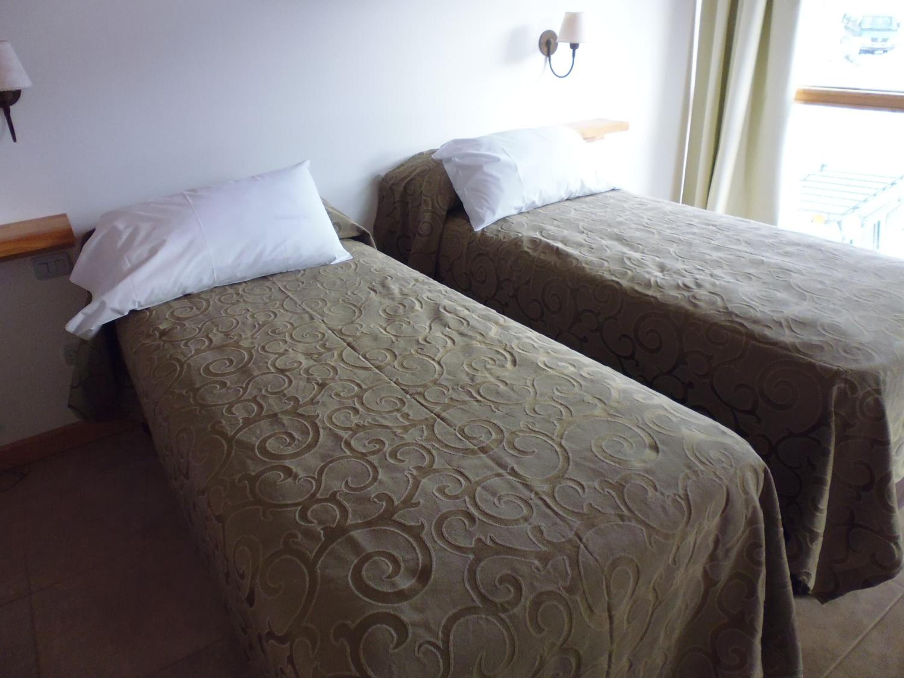 Two single beds with patterned bedspreads, a wall-mounted lamp, and a window with curtains.