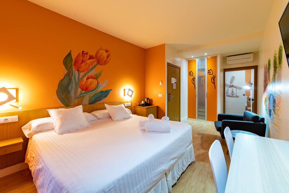 A hotel room with a large bed, a desk, and a chair. There is a painting of flowers on the wall, and a door with a window.