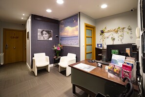 A reception area with a wooden door labeled "2 Glasgow," a desk with informational brochures, a computer, and two white chairs. - Ur-Alde (San Sebastián)